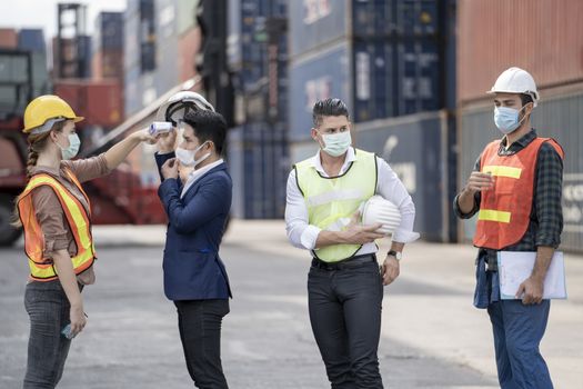 Factory woman worker in a face medical mask and safety dress used measures temperature at worker people standing on queue with a non-contact infrared thermometer.
