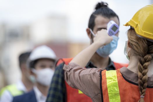 Factory woman worker in a face medical mask and safety dress used measures temperature at worker people standing on queue with a non-contact infrared thermometer.
