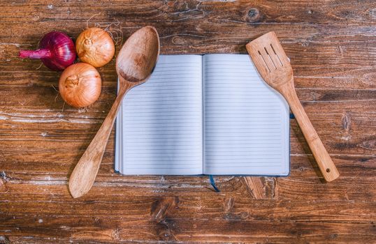 Cookbook on top of the wooden table with some onions and a wooden spoon