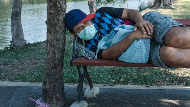 Asian homeless man wearing protective mask sleeping on a bench. Unemployed due to the impact of the spread of the virus.