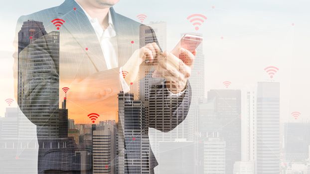 Concept of wireless connection and internet of things, IOT, shown by businessman in suit using smartphone and city as background with double exposure.