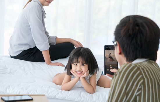 Asian family with cute parents and daughter. The father was using a cell phone to take pictures of his daughter and mother sitting nearby. They are having fun playing in the bedroom.