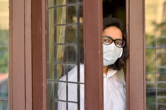 Concerned & worried lady under lock down home quarantine wearing protective breathing surgical face mask against flu and novel Corona virus (Covid-19) pandemic & greeting visitor through door/window