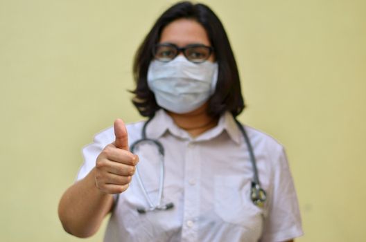 Portrait of a young medical healthcare female worker showing thumbs up sign with her right hand wearing surgical mask to protect herself from Corona Virus (COVID-19) pandemic stethoscope around neck