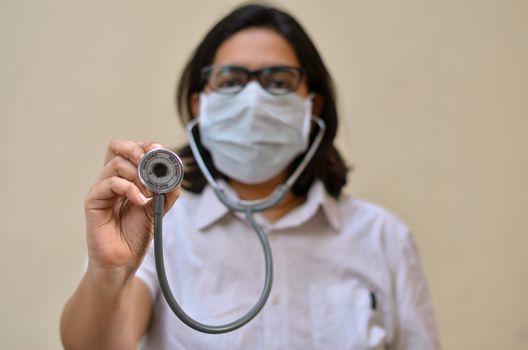 Portrait of young medical healthcare female worker showing / holding out her stethoscope & wearing surgical face mask to protect from Corona Virus (COVID-19) pandemic against yellow background