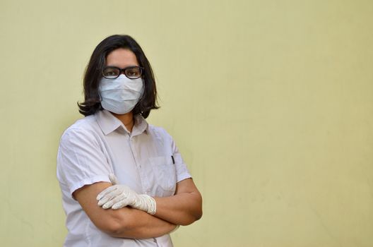 Portrait of a young medical healthcare female worker with hands crossed / folded, wearing surgical mask to protect herself from Corona Virus (COVID-19) pandemic disease against yellow background