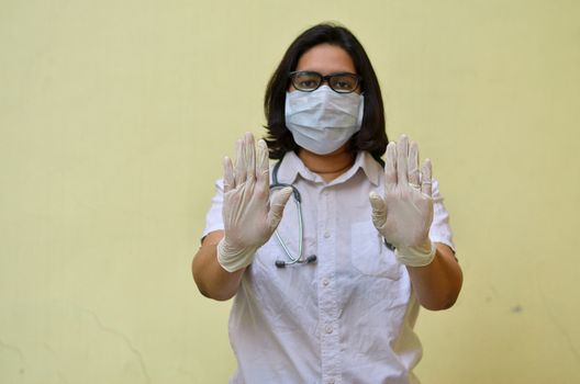 Portrait of young medical healthcare female worker showing her hands out & wearing surgical face mask to protect from Corona Virus (COVID-19) pandemic against yellow background.Concept Ready to fight