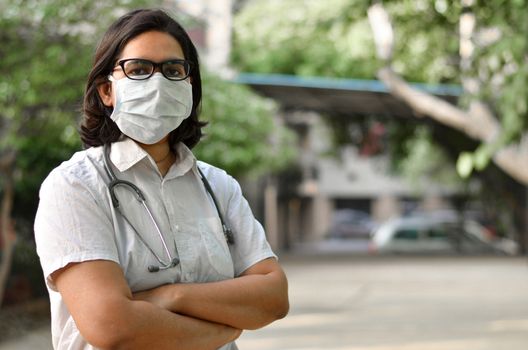 Portrait of a young medical healthcare female worker with hands crossed / folded, wearing surgical mask to protect herself from Corona Virus (COVID-19) pandemic disease against blur background