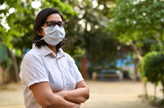 Side angle portrait of a young medical healthcare female worker with hands crossed / folded, wearing surgical mask to protect herself from Corona Virus (COVID-19) pandemic against blur background