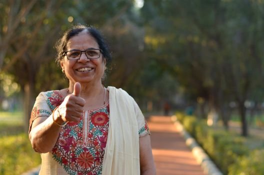 Portrait shot of a happy looking senior north Indian woman wearing traditional chikan kari Indian salwar kameez showing a thumbs up in a garden against a bokeh of canopy of trees.