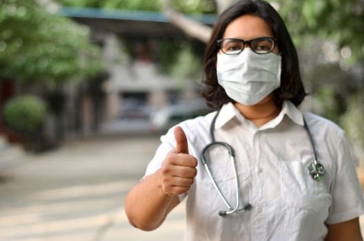 Portrait of a young medical healthcare female worker showing thumbs up sign with her right hand wearing surgical mask to protect herself from Corona Virus (COVID-19) pandemic stethoscope around neck