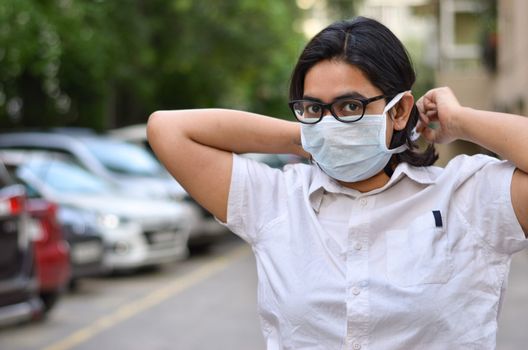 Portrait of a young medical healthcare female worker tying surgical mask on her face to protect herself from Corona Virus (COVID-19) pandemic against blur background. Concept - Getting ready to fight