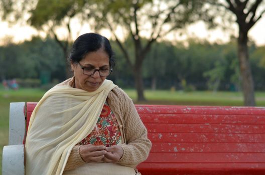 Shot of a senior retired Indian woman sitting in a park on a red bench peeling peanuts, and eating them. Concept - Retired life