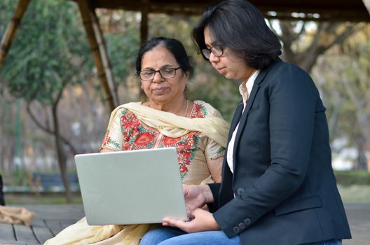 Young Indian woman helping her old retired mother on a laptop sitting in a park in New Delhi, India. Concept - Digital literacy / Education, Mother's Day
