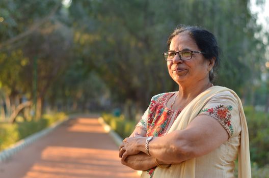 Smart and confident senior north Indian woman standing, thinking and looking away with hands crossed / folded in a park wearing an off white salwar kameez punjabi suit in summers in New Delhi, India