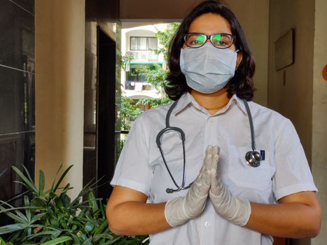 Portrait of young medical healthcare female worker hands crossed / folded in namaste, wearing surgical mask, stethoscope over shoulders to protect herself from Corona Virus (COVID-19) pandemic disease