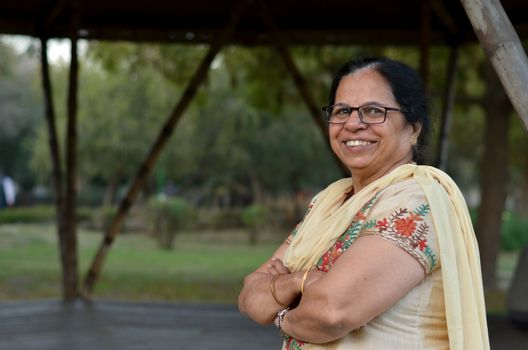 Smart and confident senior north Indian woman standing, posing for the camera with hands crossed / folded in a park wearing an off white salwar kameez punjabi suit in summers in New Delhi, India