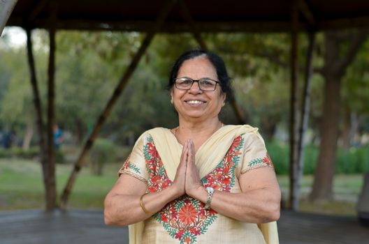 Smart senior north Indian woman standing, posing for the camera with hands folded in namaste as a sign of respect and welcome in a park wearing off white salwar kameez in summers in New Delhi, India