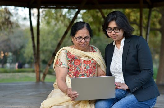 Young Indian woman helping her old retired mother on a laptop sitting in a park in New Delhi, India. Concept - Digital literacy / Education, Mother's Day