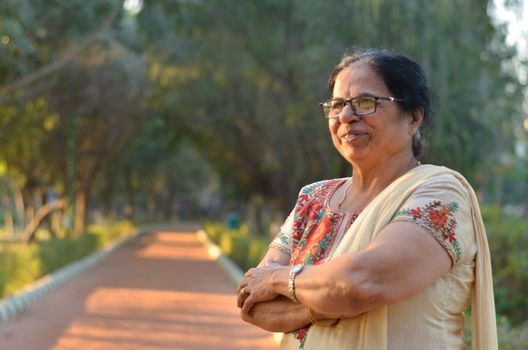 Smart and confident senior north Indian woman standing, thinking and looking away with hands crossed / folded in a park wearing an off white salwar kameez punjabi suit in summers in New Delhi, India