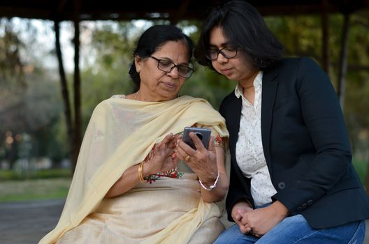 Smart young Indian girl with an old Indian woman looking at the mobile phone and busy talking sitting on a bench in a park in Delhi, India