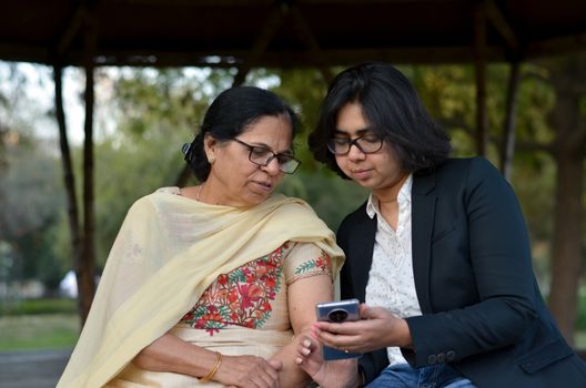 Smart young Indian girl with an old Indian woman looking at the mobile phone and busy talking sitting on a bench in a park in Delhi, India