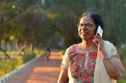 Senior Indian woman laughing and speaking on mobile smart phone in a park wearing off white salwar kamiz. Concept - Digital literacy in India for senior citizens