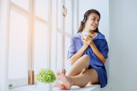 Picture of a beautiful girl who is awake, sitting, relaxing, drinking coffee, playing mobile and reading a book by the window.