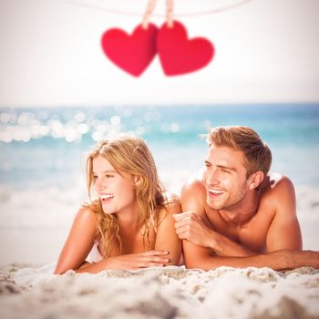 Happy couple relaxing together in the sand against hearts hanging on a line
