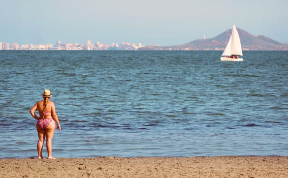 An unidentified woman standing at the seashore looking at a boat in the mediterranean sea