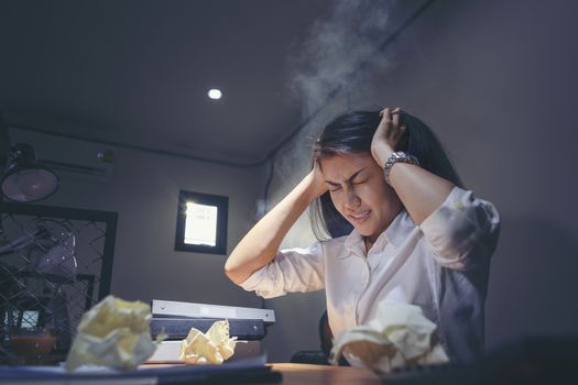 Deadline stress concept - furious business woman sitting at desk in office and shouting while holding hands on head with stress from hard work