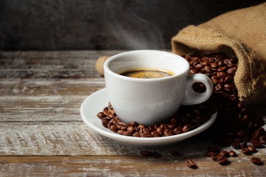 Coffee cup and roasted coffee beans in a burlap sack on a rustic wooden table on rusty vintage background.