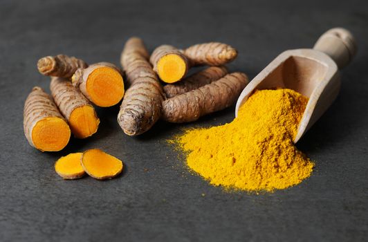 Turmeric powder healthy spice Asian food closeup of turmeric roots sliced and a wooden bailer on a rustic dark grey kitchen board.