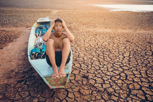 Sad a boy sitting on a boat that was parked in an arid ground for the hope of the sky to rain. Affected of global warming made climate change. Water shortage and drought concept.