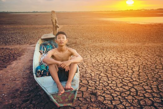 Sad a boy sitting on a boat that was parked in an arid ground for the hope of the sky to rain. Affected of global warming made climate change. Water shortage and drought concept.