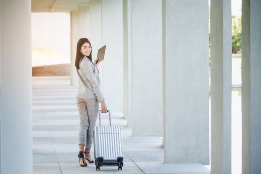 Portrait of business woman looking digital tablet with white travel bag on walkway while waiting to travel to the destination 