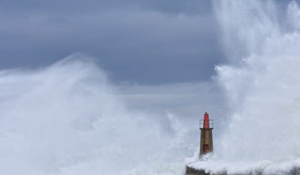 Stormy wave over old lighthouse and pier of Viavelez in Asturias, Spain.