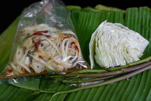 Thai food called papaya salad, pickled fish, salted eggs in a plastic bag and vegetables on a banana leaf, green, black background