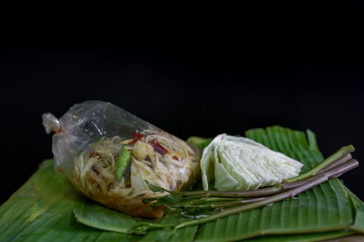 Thai food called papaya salad, pickled fish, salted eggs in a plastic bag and vegetables on a banana leaf, green, black background