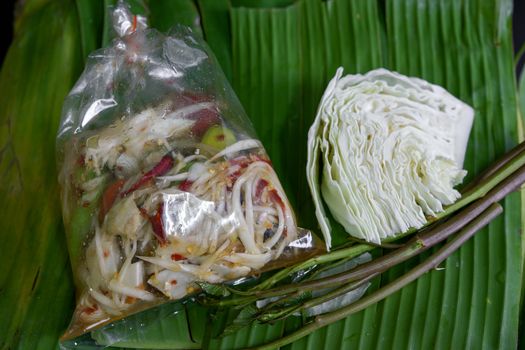Thai food called papaya salad, pickled fish, salted eggs in a plastic bag and vegetables on a banana leaf, green, black background