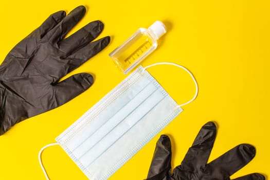 Black gloves with blue mask on yellow background with homemade hand sanitizer gel in plastic bottle.