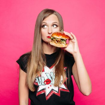 Front view of sadly young girl looking at side and thinking eating tasty humburger or no. Pretty woman wearing in black t-shirt holding big cheeseburger, posing on pink background. Concept of diet.