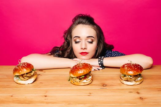 Portrait of unhappy pretty girl with hairstyle and make up looking at delicious freshly cooked hamburgers on wooden table with her chin on hands over bright magenta background.