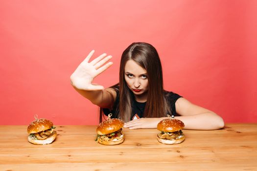 Studio portrait of beautiful young woman with long straight hair holding her hand at camera with stop gesture protesting against unhealthy burgers on wooden table in front of her. Isolate on red background.