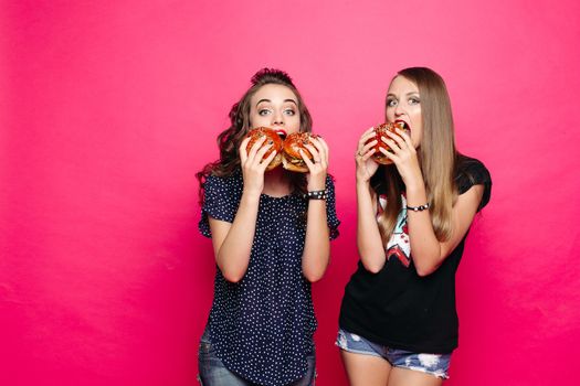 Studio portrait of girlfriends eating delicious and yummy burgers with chicken with hungry look at camera against vivid magenta background.