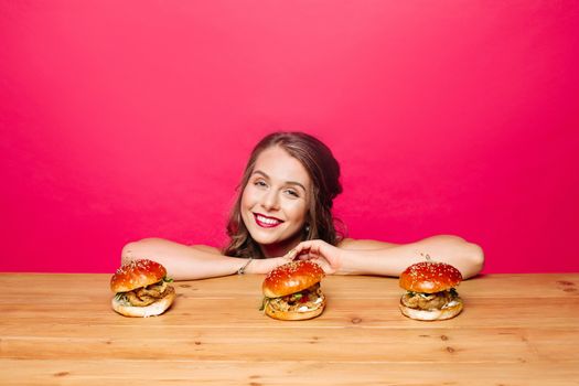 Portrait of beautiful happy girl smiling at camera sitting in front of three yummy burgers on wooden table against pink background.