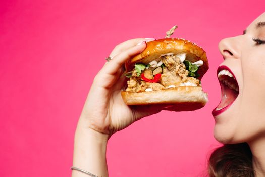 Woman eating sandwich. The happy girl is eating a hamburger. She opened her mouth, holding a hamburger in her right hand and looking at it appetizingly. A student with a meal on a pink background