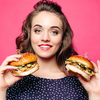 Beautiful positivity girl smilingat camera and posing with two tasty humburgers. Young pretty woman wearing in black holding big cheeseburgers. Pink studio background. Concept of fast food and diet.