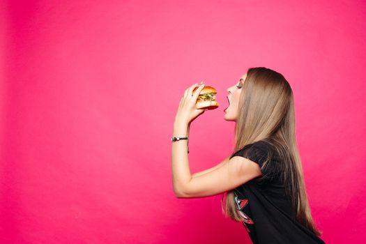 Close-up of pretty long-haired girl biting delicious burger with chicken and salad, looking at camera against pink background. American fast food concept.