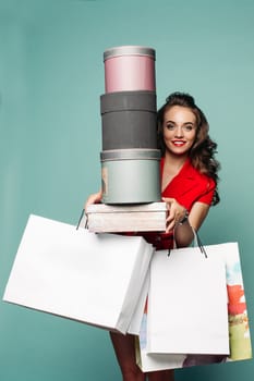 Studio portrait of trendy pretty girl with wavy hair in red smiling at camera holding boxes and shopping bags after shopping in mall over green background. Smiling at camera holding new clothes and shoes with hats.